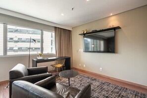 A modern living room with a sofa, a small round table, a chair, a TV mounted on the wall, and a large window with curtains. - #RD1109 | Cyclinn Vila Olímpia (Vila Olímpia)