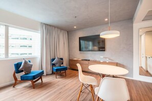 Une pièce moderne comprenant une table à manger, deux chaises, un canapé, un téléviseur fixé au mur et une grande fenêtre avec des rideaux.