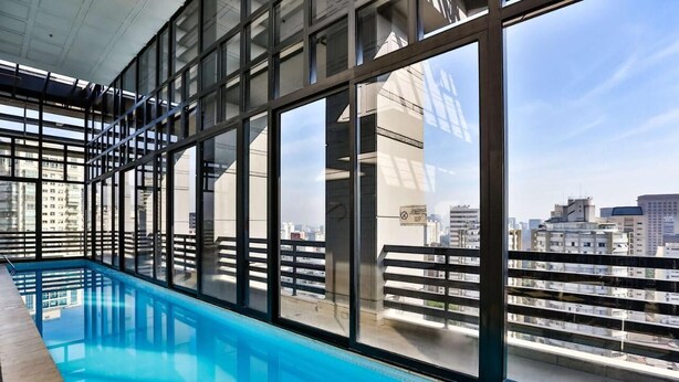 An indoor swimming pool with large windows offering city views. - #RB1107 | Cyclinn Cidade Jardim Itaim (Itaim Bibi)