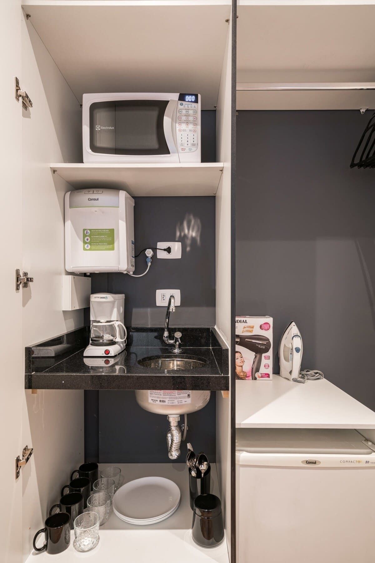 A compact kitchenette with a microwave, coffee maker, sink, and storage for mugs and glasses.