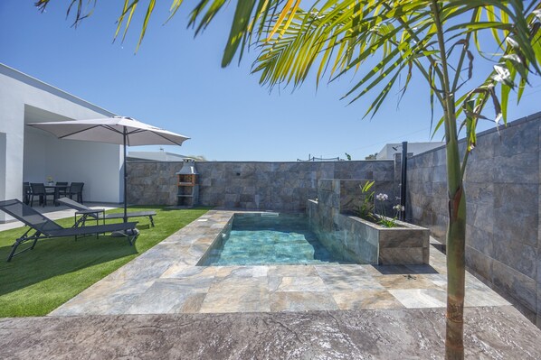Une piscine bordée de carreaux de pierre, une pelouse et une terrasse avec des chaises et un parasol.