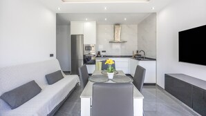 Un salon moderne avec un canapé, une table à manger, des chaises, un téléviseur et une cuisine en arrière-plan.