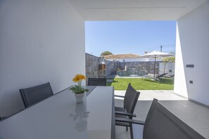 Une cuisine moderne dotée d’un grand îlot central, d’un coin repas avec une table et des chaises, et offrant une vue sur un jardin avec une piscine et un espace salon extérieur.