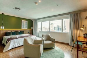 Ein Hotelzimmer mit Bett, zwei Sesseln, einem Esstisch, Blick auf die Stadt und einem großen Fenster.