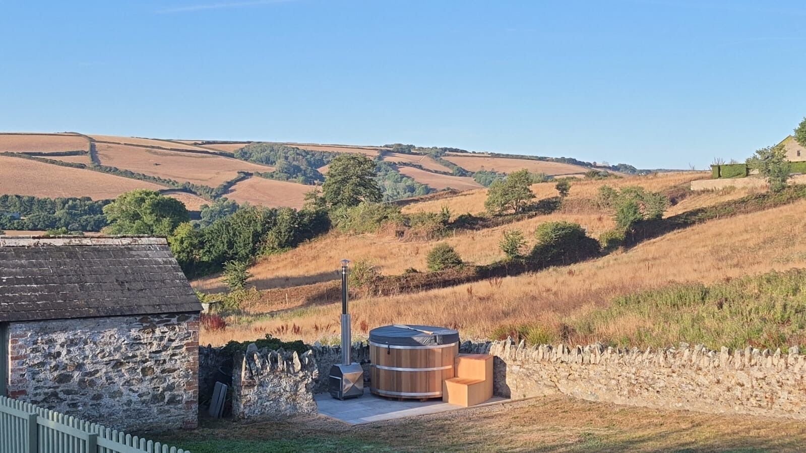 A rustic stone building with a chimney, a hot tub, and a stone wall in a rural landscape.