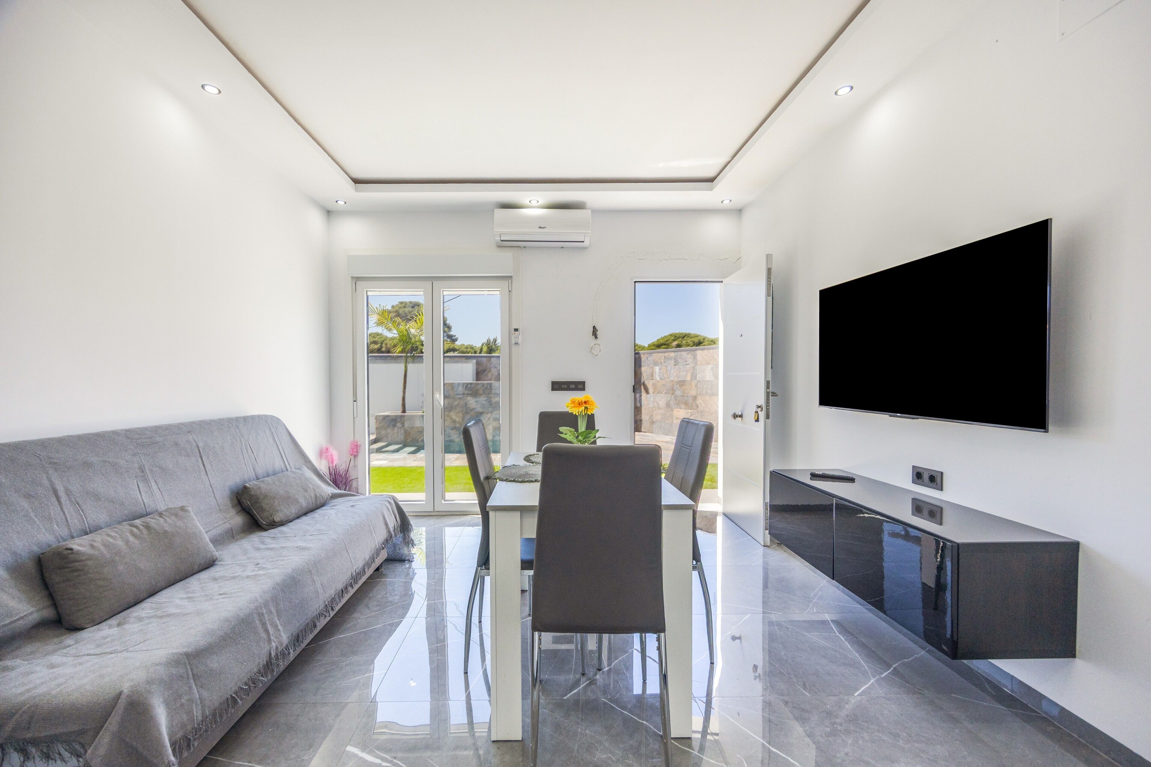 A modern living room with a sofa, dining table, chairs, a flat-screen TV, and a kitchen area.