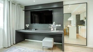 Una camera moderna con una scrivania grigio scuro, una grande TV a schermo piatto, una piccola panca bianca e una zona cucina visibile attraverso una parete divisoria in vetro.