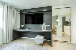 Ein modernes Zimmer mit einem dunkelgrauen Schreibtisch, einem großen Flachbildfernseher, einer kleinen weißen Bank und einer durch eine Glaswand abgetrennten Küchenzeile.