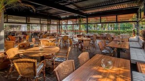 Restaurante interno com várias mesas e cadeiras, grandes janelas e vista para paisagem tropical.