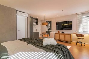 A modern bedroom with a bed, a desk, a chair, a TV, and a kitchenette. - #TR1006 | Cyclinn Faria Lima Itaim (Itaim Bibi)