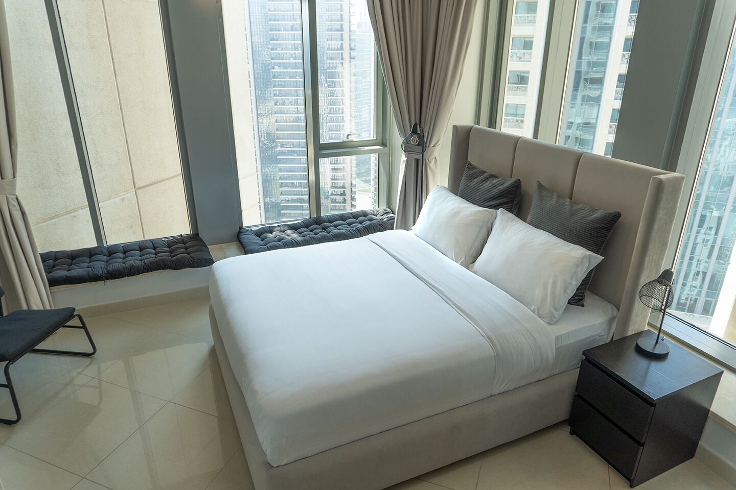 Une chambre moderne avec un grand lit, une table de chevet et une vue sur de hauts immeubles, visible à travers de grandes fenêtres.