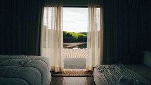 A hotel room with two beds, a view of a green landscape through sheer curtains, and a wooden deck outside.