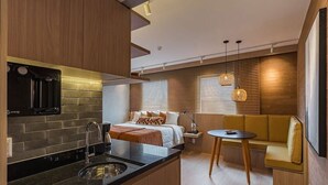 A compact hotel room with a kitchenette, a bed with a nightstand, a seating area with a table, and a large window with blinds. - #BE1902 | Cyclinn Vila Olímpia Berrini (Berrini)