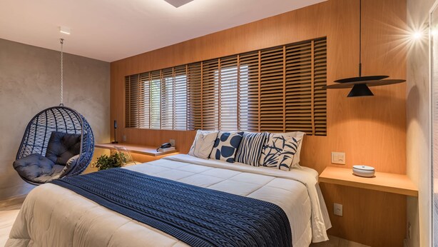 Ein modernes Schlafzimmer mit einem großen Bett, einem Hängekorbsessel, Lamellen an der Fensterverkleidung und einem Nachttisch mit Lampe.