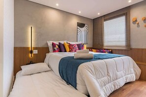 A modern bedroom with a large bed, a bedside table, a wall-mounted light, and a window with blinds. - #QL1301 | Cyclinn Faria Lima Pinheiros (Pinheiros)