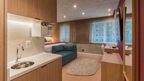 Ein modernes Zimmer mit integrierter Küche, einer Couch, einem runden Teppich und großen Fenstern mit Jalousien.