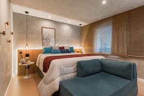 A modern bedroom with a bed, a sofa, a bedside table, and a window with blinds. - #BE601 | Cyclinn Vila Olímpia Berrini (Berrini)
