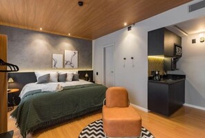 A modern bedroom with a bed, a small seating area, a kitchenette, and a door. - #IN1704 | Cyclinn Jesuíno Arruda Itaim (Itaim Bibi)