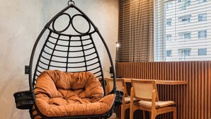 Ein Zimmer mit Hängesessel, Holztisch und Blick auf ein Gebäude durch Jalousien.