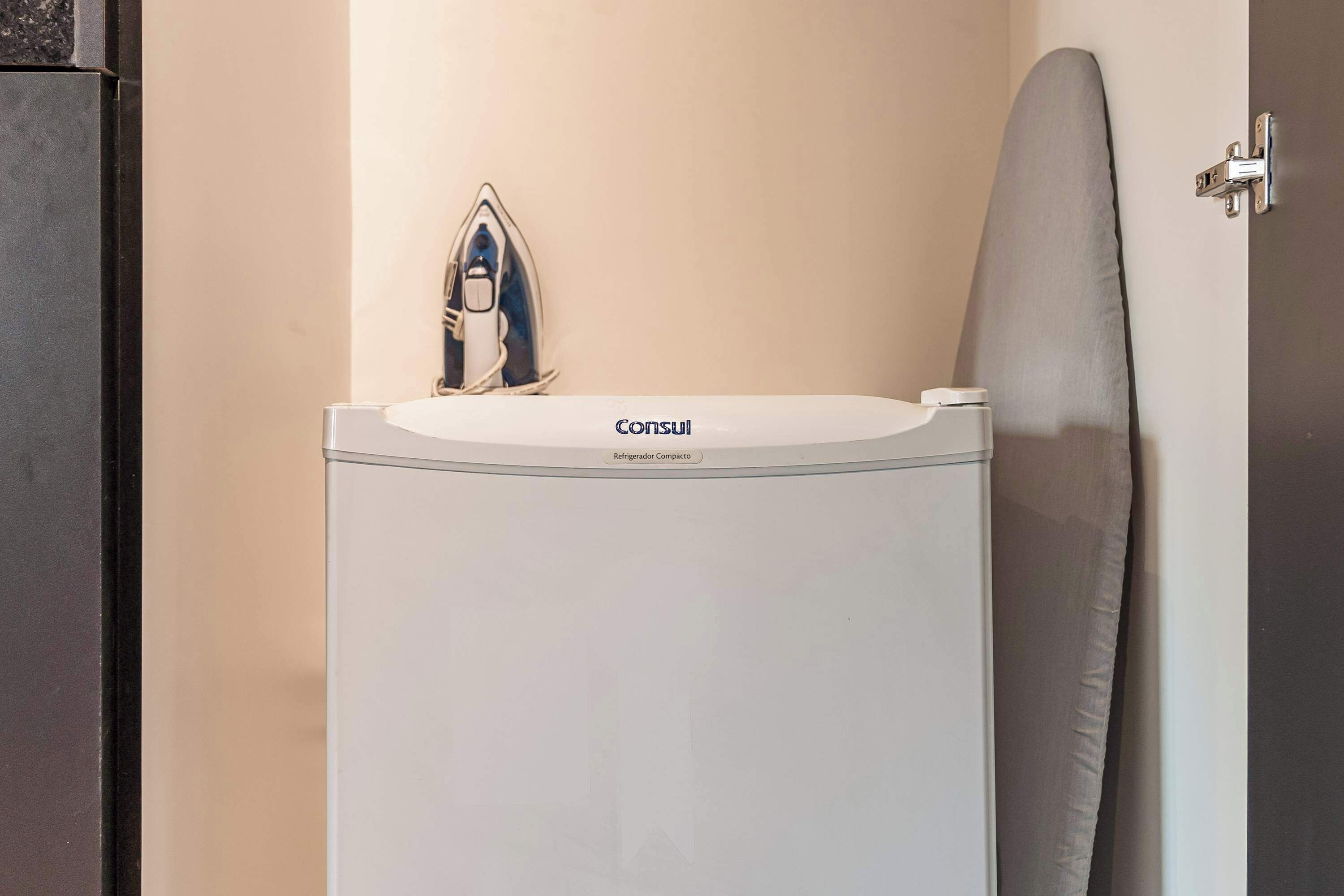 A compact refrigerator with a built-in iron on top, a grey ironing board, and a door with a silver handle.