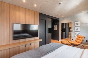 Ein modernes Schlafzimmer mit einem großen Flachbildfernseher, einem Bett mit grauer Decke, einem Ledersessel und einer Wand aus Holzpaneelen.
