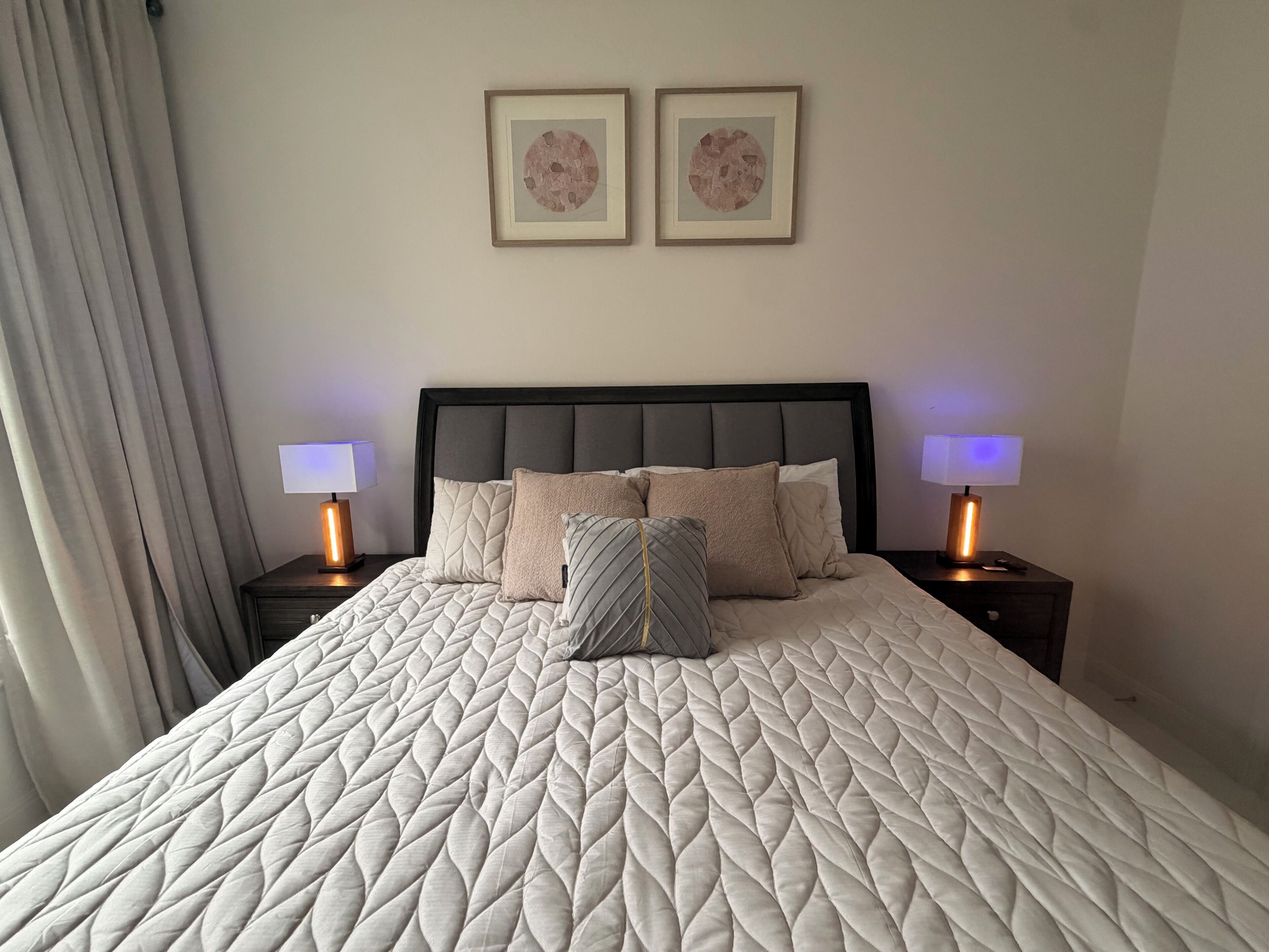 A neatly made bed with a quilted headboard, two bedside lamps, and two framed artworks on the wall.
