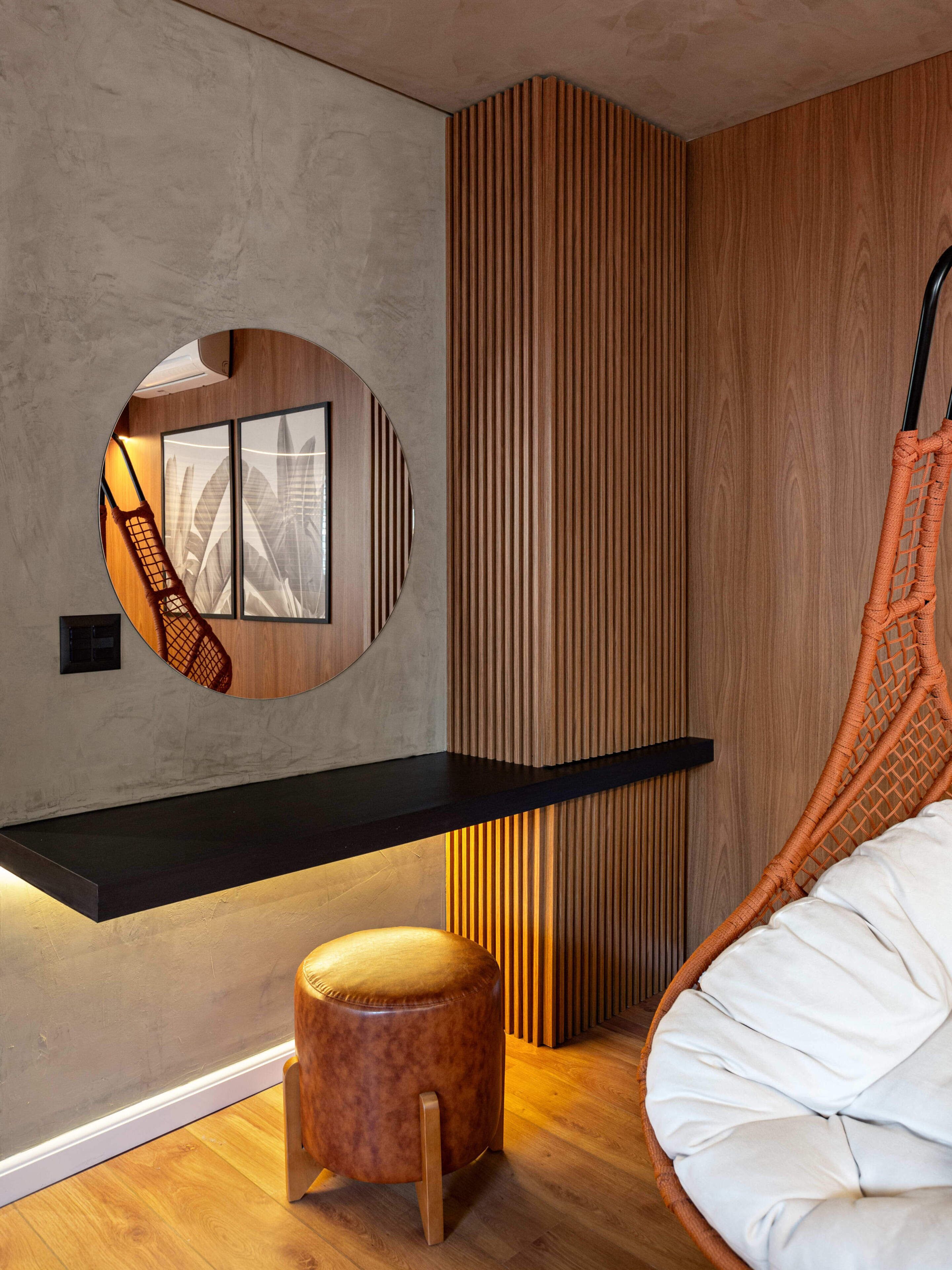 Una habitación moderna con un espejo circular, un estante negro, un revestimiento de pared de listones de madera, una hamaca colgante y un otomano redondo.