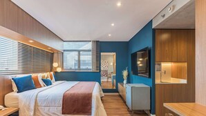 Une chambre à coucher moderne, avec un lit, des tables de chevet, un bureau, une télévision et une salle de bain visible à travers une porte.