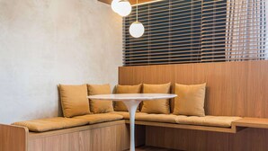 Un coin salon avec des sièges disposés en angle, une table ronde blanche, un lambris en bois et une fenêtre avec des stores.
