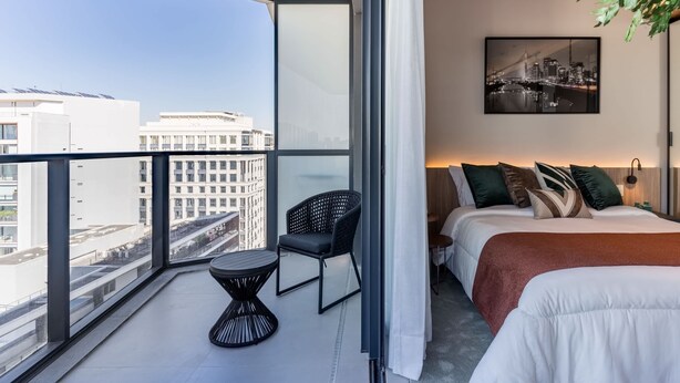 A modern hotel room with a balcony, a bed with multiple pillows, a small table, and a cityscape view. - #VN1811 | Cyclinn Vila Olímpia (Vila Olímpia)