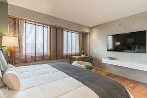 A modern bedroom with a large bed, a desk, a chair, a TV, and large windows with blinds. - #QL0603 | Cyclinn Faria Lima Pinheiros (Pinheiros)