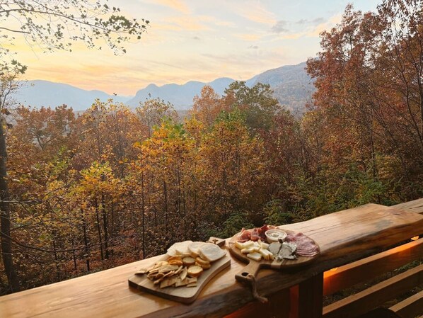 Un banc en bois, sur lequel sont disposés divers aliments, offre une vue sur un paysage boisé avec des montagnes à l’horizon.