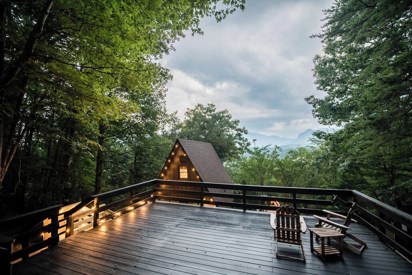 A wooden cabin with a deck, surrounded by trees and a mountain view.