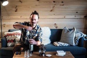 Eine Person sitzt auf einem Sofa, auf dem Tisch stehen eine Flasche, Gläser und eine Fernbedienung.