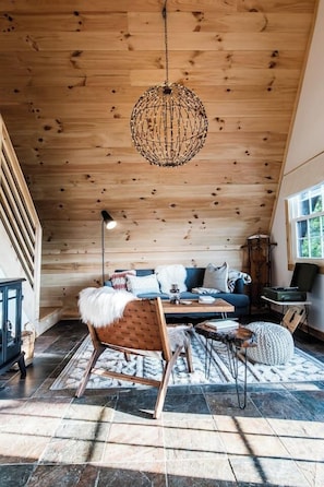 A cozy cabin interior with a wooden ceiling, a hanging light fixture, a sofa with cushions, a rocking chair, and a small table. - Nordic A-Frame Getaway: Hot Tub·EpicView·Secluded (Lake Lure)