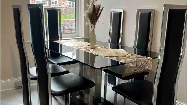 Een eethoek met een glazen tafel, zwarte stoelen en een vaas met gedroogde bloemen.