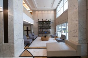 Um lobby moderno com piso de mármore, área de estar central com sofás, uma estante e janelas amplas.