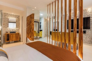 Une chambre d’hôtel moderne avec un lit, une cloison en bois, un mur en pierre et un coin repas avec des chaises jaunes.