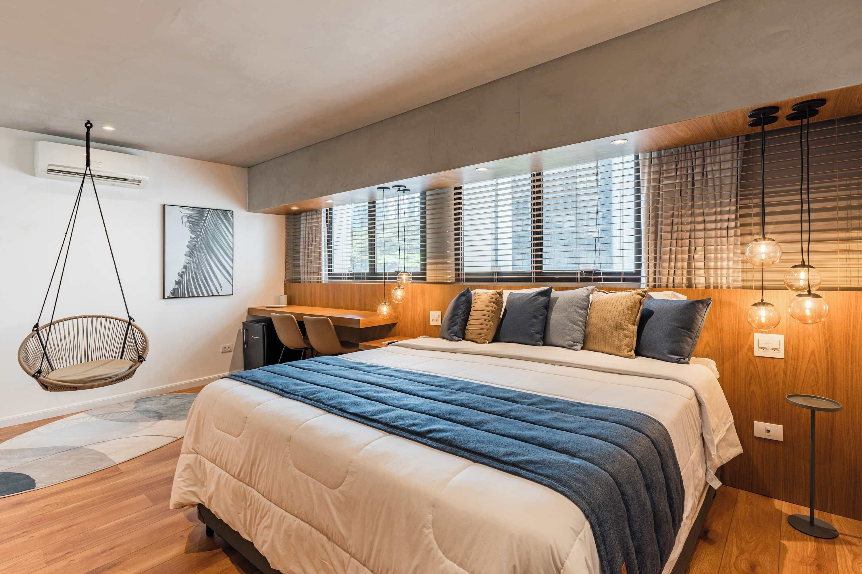 A modern bedroom with a large bed, a hanging chair, a desk, and a window with blinds.