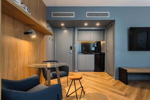 Ein modernes Zimmer mit einer Holzwand, einem runden Glastisch, zwei blauen Stühlen, einem Hocker, einer Bank und einer Küchenzeile mit grauen Schränken und einem schwarzen Kühlschrank.