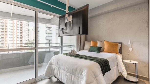Et moderne hotelværelse med en stor seng, et tv ophængt på væggen, en balkon med udsigt til høje bygninger og et lille sengebord.