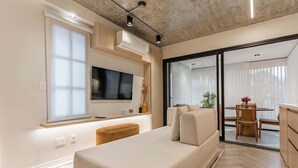 Uma sala de estar moderna com sofá, TV de tela plana, área de jantar com mesa e cadeiras, e uma porta de vidro deslizante para uma varanda.