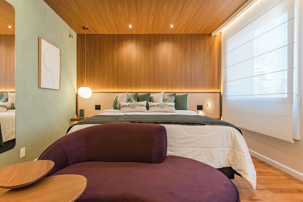 Ein modernes Schlafzimmer mit Holzbalkendecke, einem Bett mit grün-weißer Bettwäsche, einem violetten Sessel und einem Nachttisch.