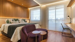Une chambre moderne avec un lit, un fauteuil violet, une petite table ronde et une vue sur la ville à travers de grandes fenêtres.