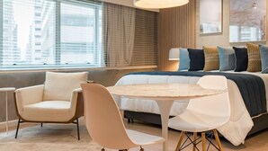 Un hotel moderno con cama, dos sillas, una mesita redonda, una lámpara colgante grande y vistas de la ciudad a través de amplias ventanas.