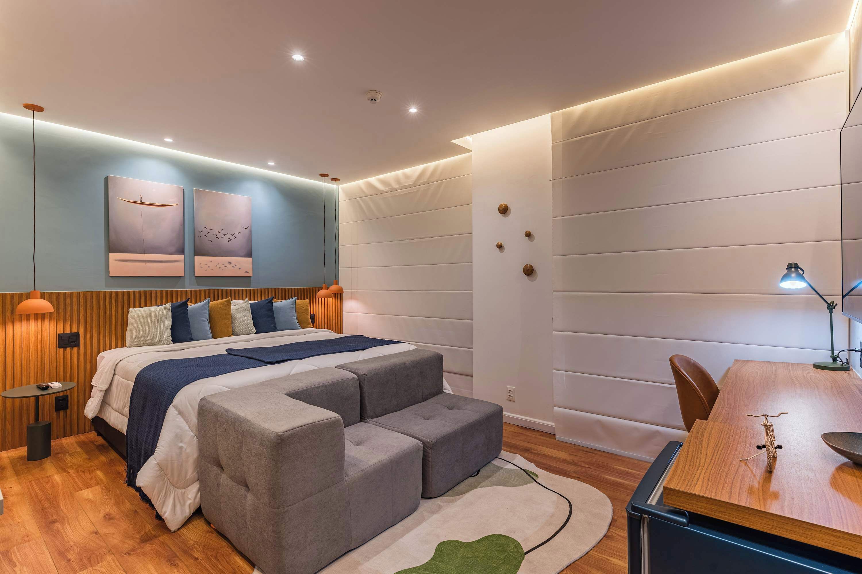 Ein modernes Schlafzimmer mit Bett, grauen Hocker, einem Holztisch und Wandbildern.