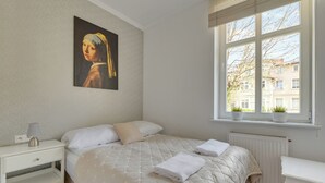 A bedroom with a bed, bedside table, lamp, window with view, radiator, and artwork on the wall. - Nadmorski Zakątek by Grand Apartments (Sopot)