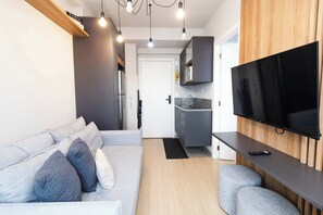 A modern living room with a sofa, a flat-screen TV mounted on a wooden wall, and a kitchen area in the background. - Studio Completo na Av. Santo Amaro | NI1802 (Vila Nova Conceição)