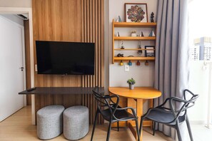 A modern living space with a flat-screen TV mounted on a wooden wall, a wooden dining table with two chairs, a small round table with a potted plant, and a shelf with decorative items. - Studio Completo na Av. Santo Amaro | NI1802 (Vila Nova Conceição)