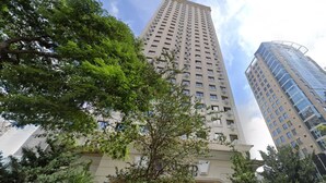 Un edificio alto de varios pisos, rodeado de árboles y otro edificio moderno.